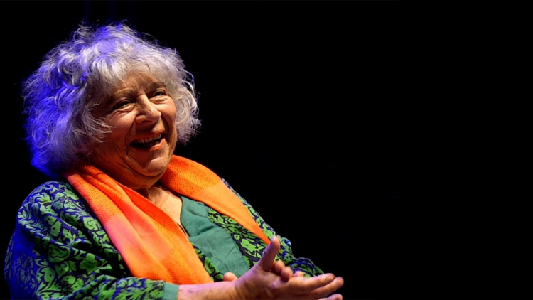 Miriam Margolyes speaks on stage wearing a bright orange scarf and green top