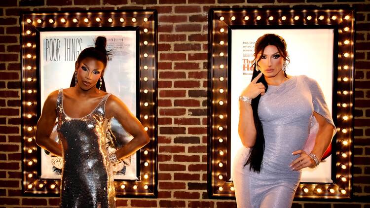 Two drag queens in front of a sign