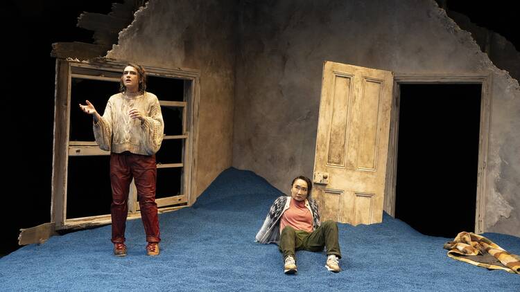 Two actors on a stage covered in blue sand, in front of a decaying house. 