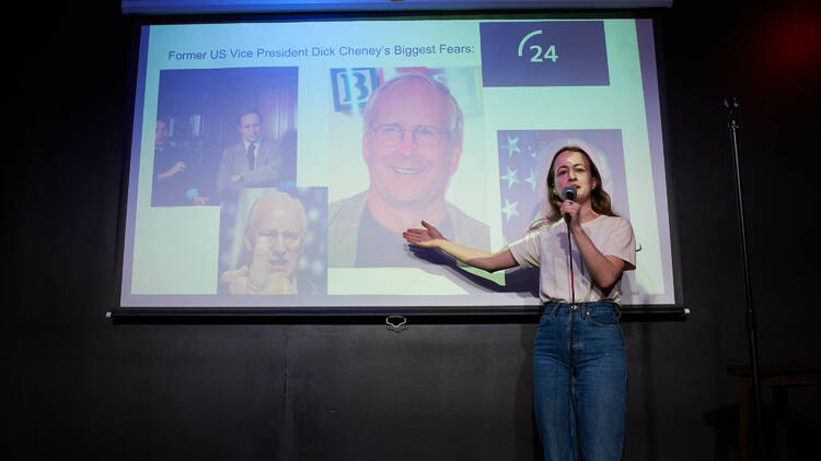 A comedian stands on stage in front of a PowerPoint slide.