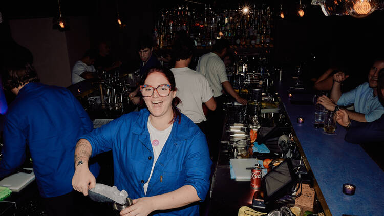 A woman in a blue long-sleeved shirt serving a drink and smiling.