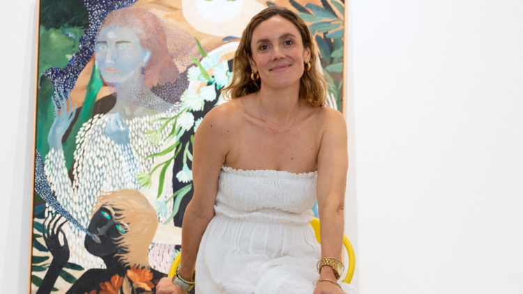 An artist wearing white clothes, sitting in front of one of her paintings