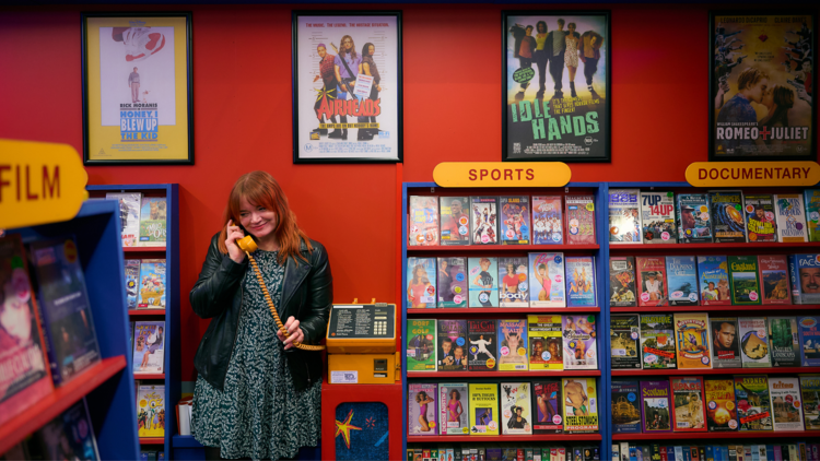 A woman holds a yellow corded phone in a replica nineties video store