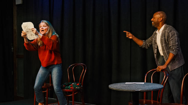 Two actors on stage, with one holding a paper and the other pointing at her.