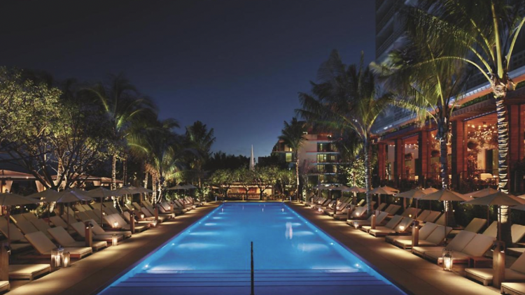 Illuminated pool at nigh time  surrounded by row of deck chairs and palm trees