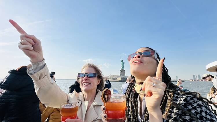 Two women stand on the deck of a boat holding cocktails while wearing eclipse glasses.