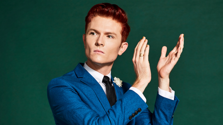 Rhys looks to the side with hands raised next to their face, wearing a blue suit