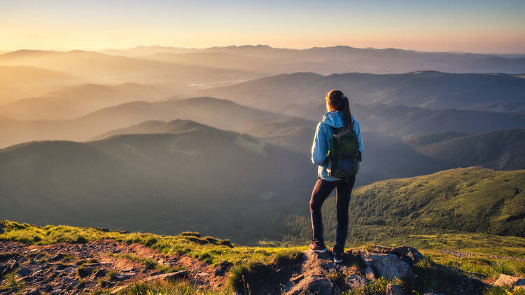 Girl on mountain peak with green grass looking at beautiful mountain valley in fog at sunset