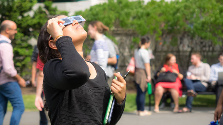  A young woman standing on a lawn and observing a partial solar eclipse using protective solar eclipse glasses.