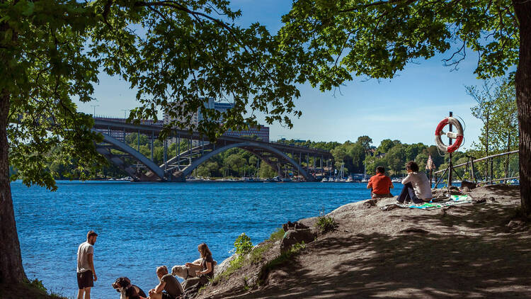 Stockholm, Sweden, June 6, 2017: View of strand on Langholmen island. Backside of people, dogs looking at city, sitting on stones, relaxing. Vasterbron bridge on background. Bright sun making shadows.