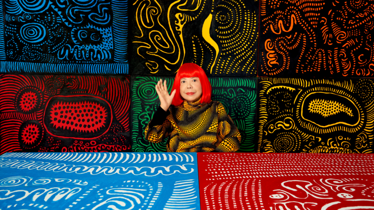 Yayoi Kusama with red hair in front of her artworks, wearing a matching sweater