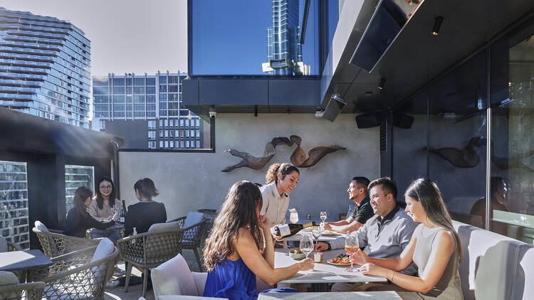 Guests enjoying the wrap-around terrace at a sunny rooftop bar with city views.