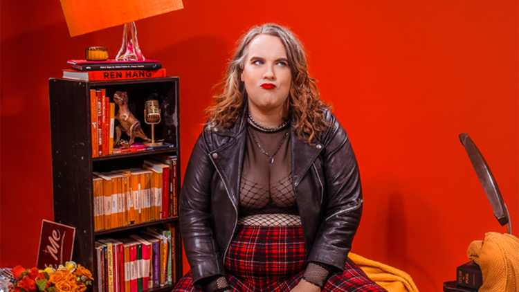 Anna wears a mesh top and red tartan pants, sitting pouting in front of a book shelf