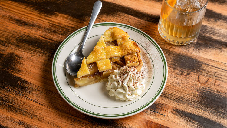 Plate of apple pie with cream and a cocktail on a wooden table.