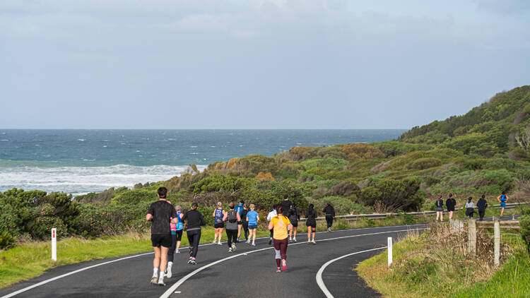 People running along the road with the ocean in the background. 