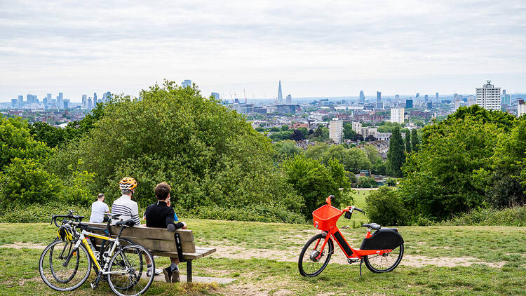 Cyclists sit on a park bench with their bikes looking at a view of the London skyline