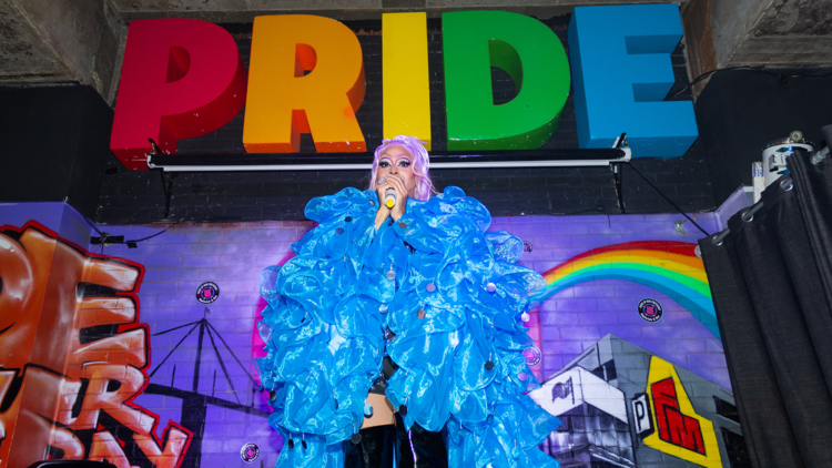 A drag queen in a big blue cape on a stage under rainbow pride letters