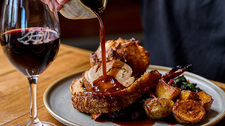 Diner pouring gravy on a Sunday roast dish with a glass of red wine.