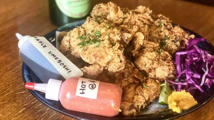 A plate of B.K. fried chicken with hand-labeled bottles of hot sauce sits on a table at Brassica Kitchen, next to a bottle and a glass of white wine