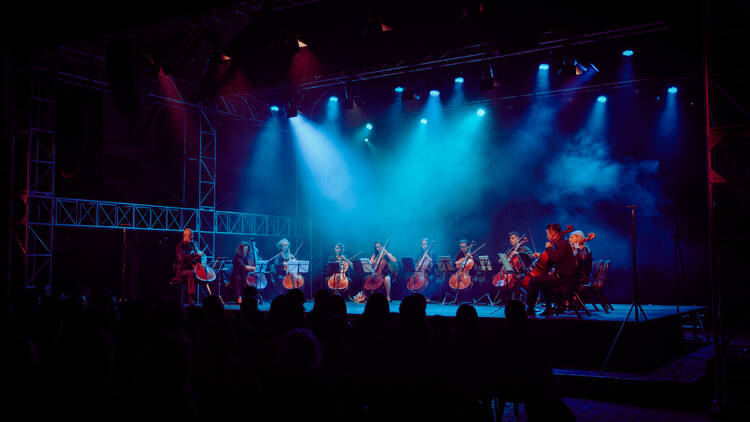 A group of cello players performing on a blue-lit stage.