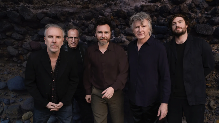 The band Crowded House standing in front of a rocky stone wall.