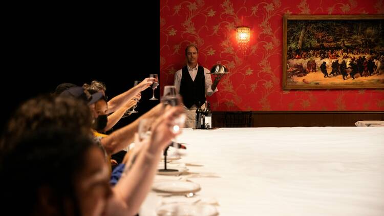 A waiter stands near a red wall while people at a dinner table hold up their glasses.