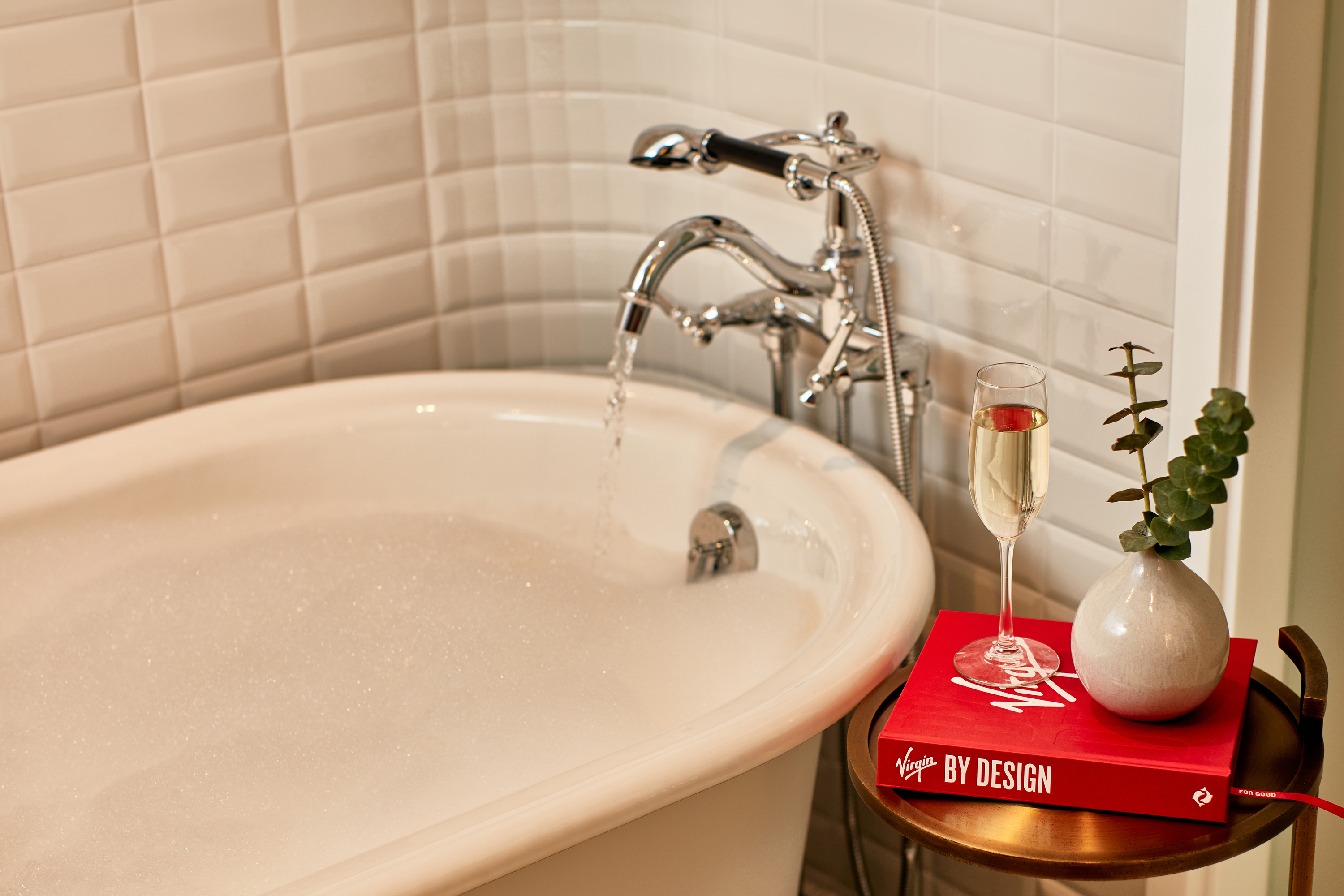 White bath tub filled with running water and bubbles with red book and glass of Prosecco to its right