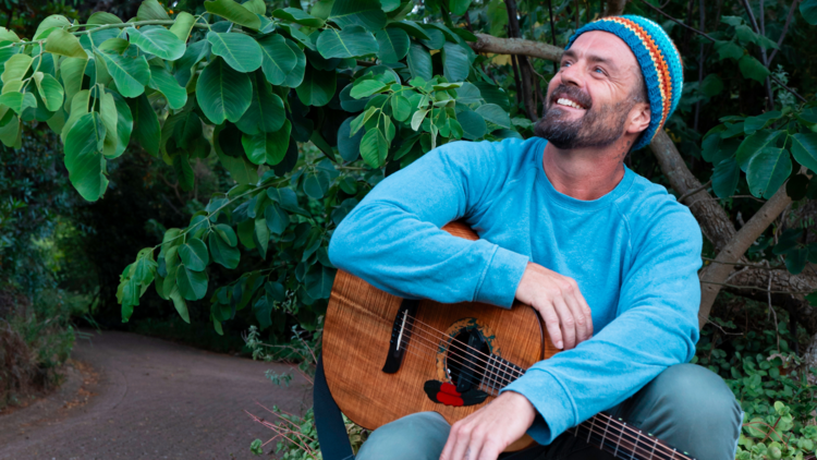 Xavier Rudd smiles wearing a bright blue casual jumper, a multi-coloured beanie and holding a guitar