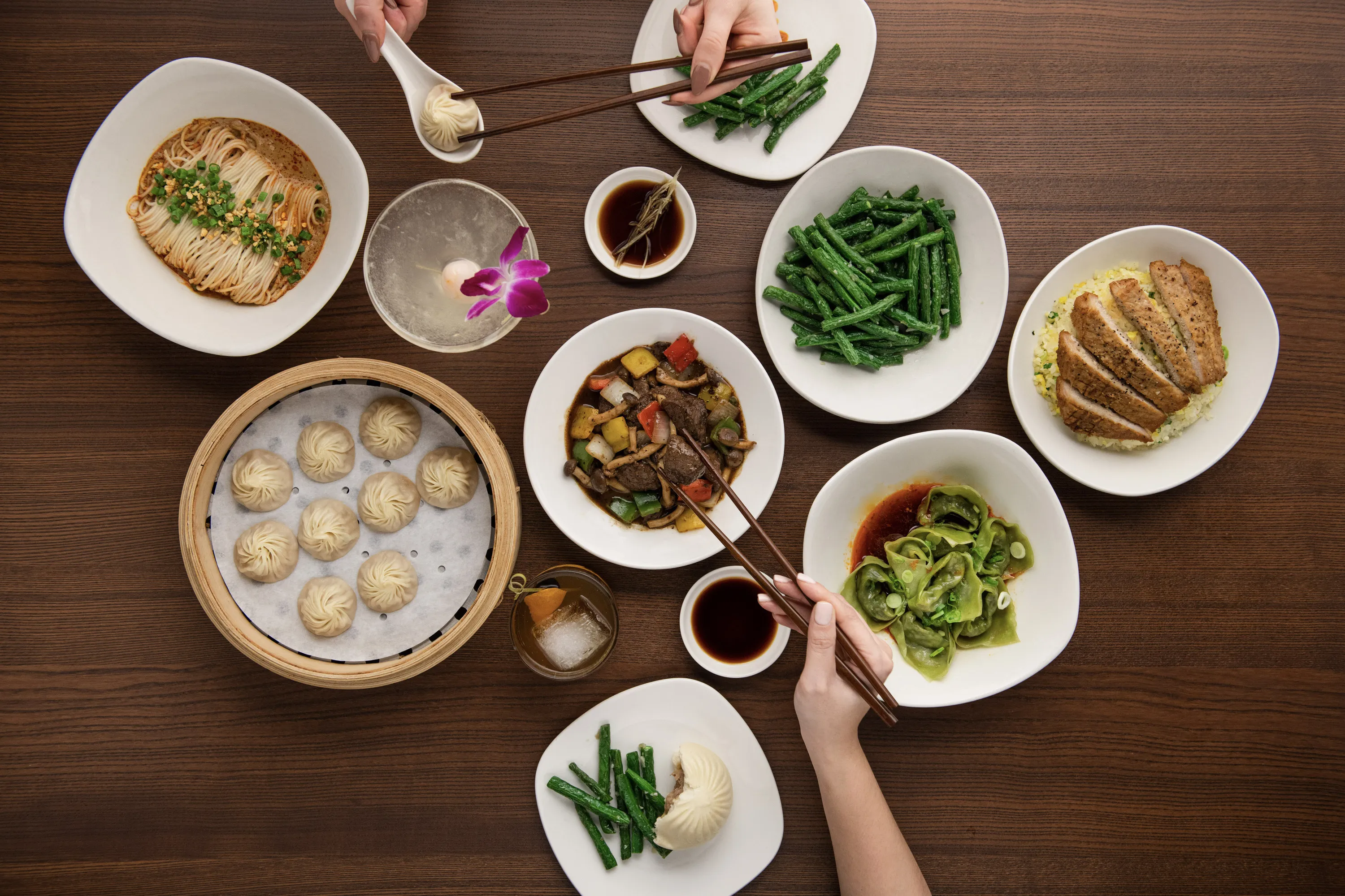Plates of xiao long bao, noodles, green beans and on a wooden table. Two hands with chopsticks dig in. 