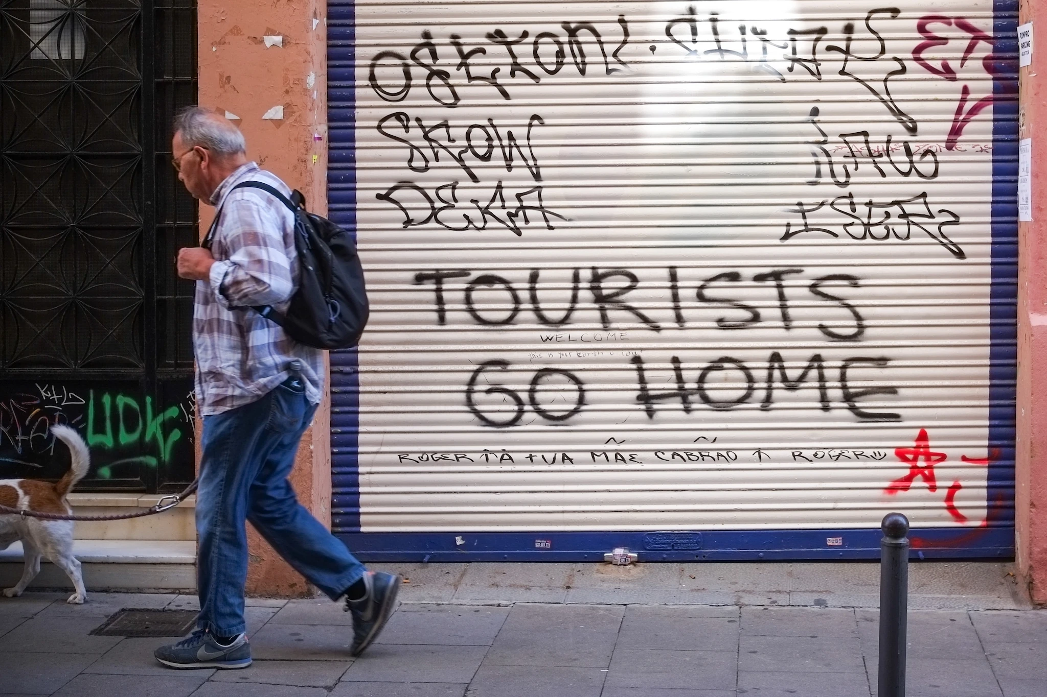 Barcelona, Spain: A man walks down the street, passing by a graffiti on a closed shop shutter that reads "Tourists go home"