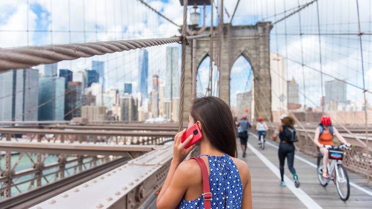 Woman using phone while walking on Brooklyn Bridge towards Manhattan city skyline. 