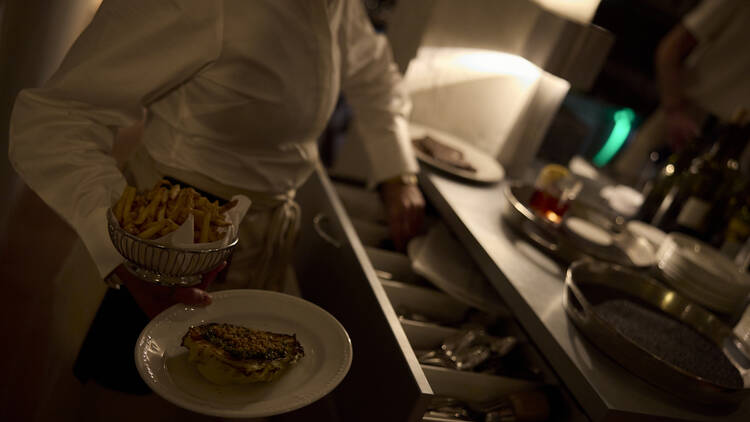 Waitress carrying a basket of chips and another dish whilst selecting cutlery from an open drawer.