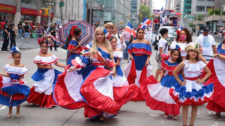 A group of women and girls in red, white, and blue dresses dance at the Dominican Day Parade.