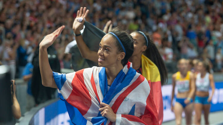 Katarina Johnson-Thompson waves to spectators whilst wearing the British flag on an athletics track. 