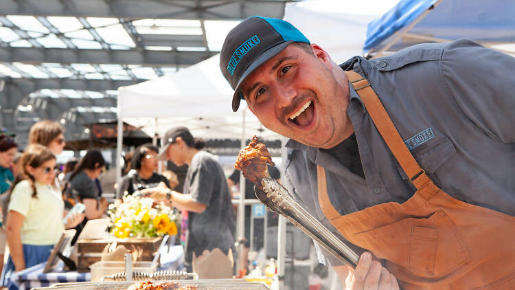 A chef in a yellow apron holding a chicken wing with a tong