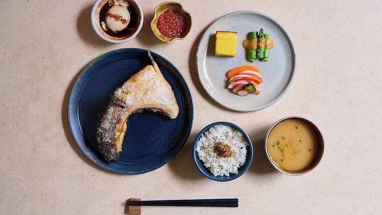 Japanese breakfast layout including grilled fish collar, sides, miso soup, rice and an onsen egg with a set of chopstick underneath