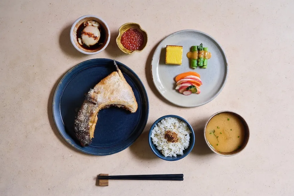 Japanese breakfast layout including grilled fish collar, sides, miso soup, rice and an onsen egg with a set of chopstick underneath