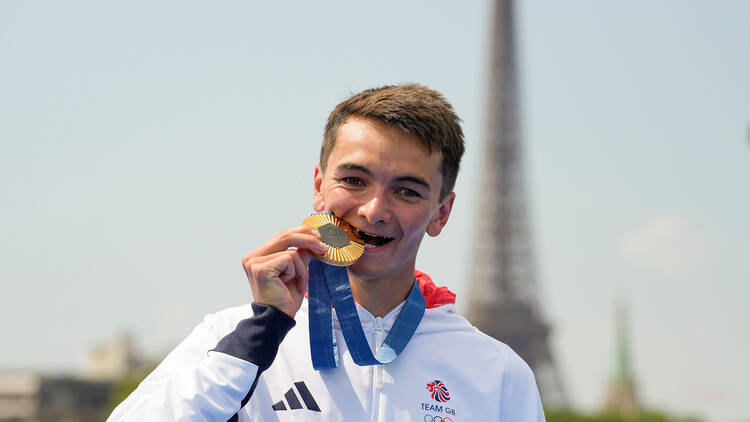 Triathlete Alex Yee biting his gold medal. Yee wears a white hoodie, with Great Britain written on it in red. The Eiffel Tower is in the background. 