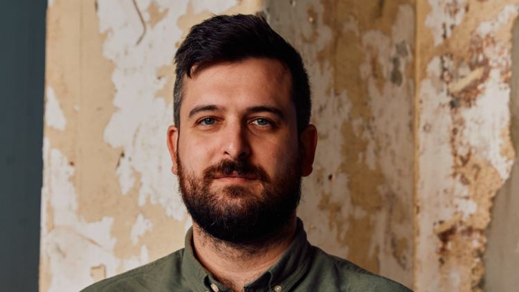 A man with a beard and moustache standing against a wall with peeling paint./