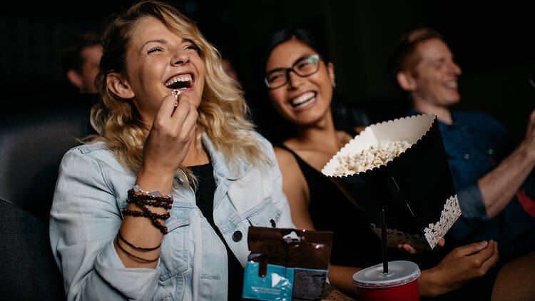 Young woman with friends watching movie in cinema and laughing. Group of people in theatre with popcorns and drinks