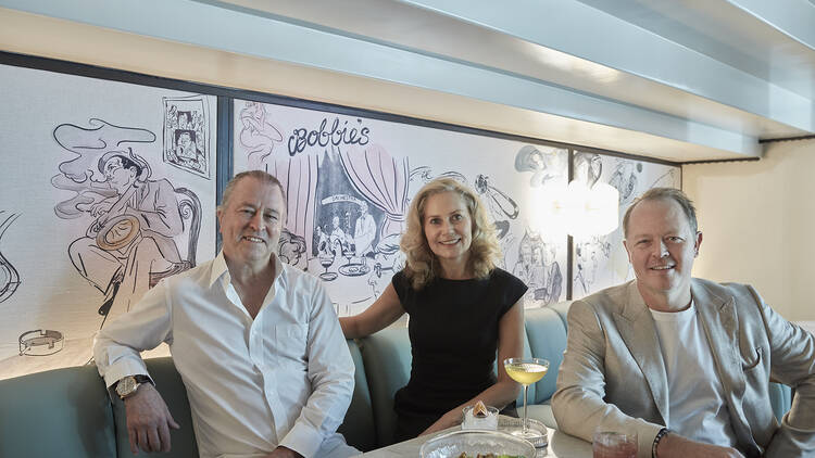 Three people sitting in a bar with an illustration on the wall and the word, 'Bobbie's'