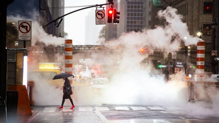 A woman with an umbrella and red high heels shoes is crossing the 42nd street in Manhattan. Cars and steam coming out from from the manholes in the background. New York City, Usa.