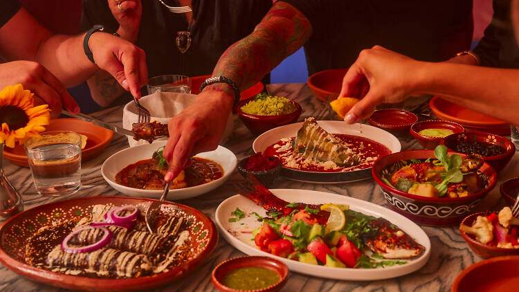 Table full of enchiladas, whole fish and people eating it with forks