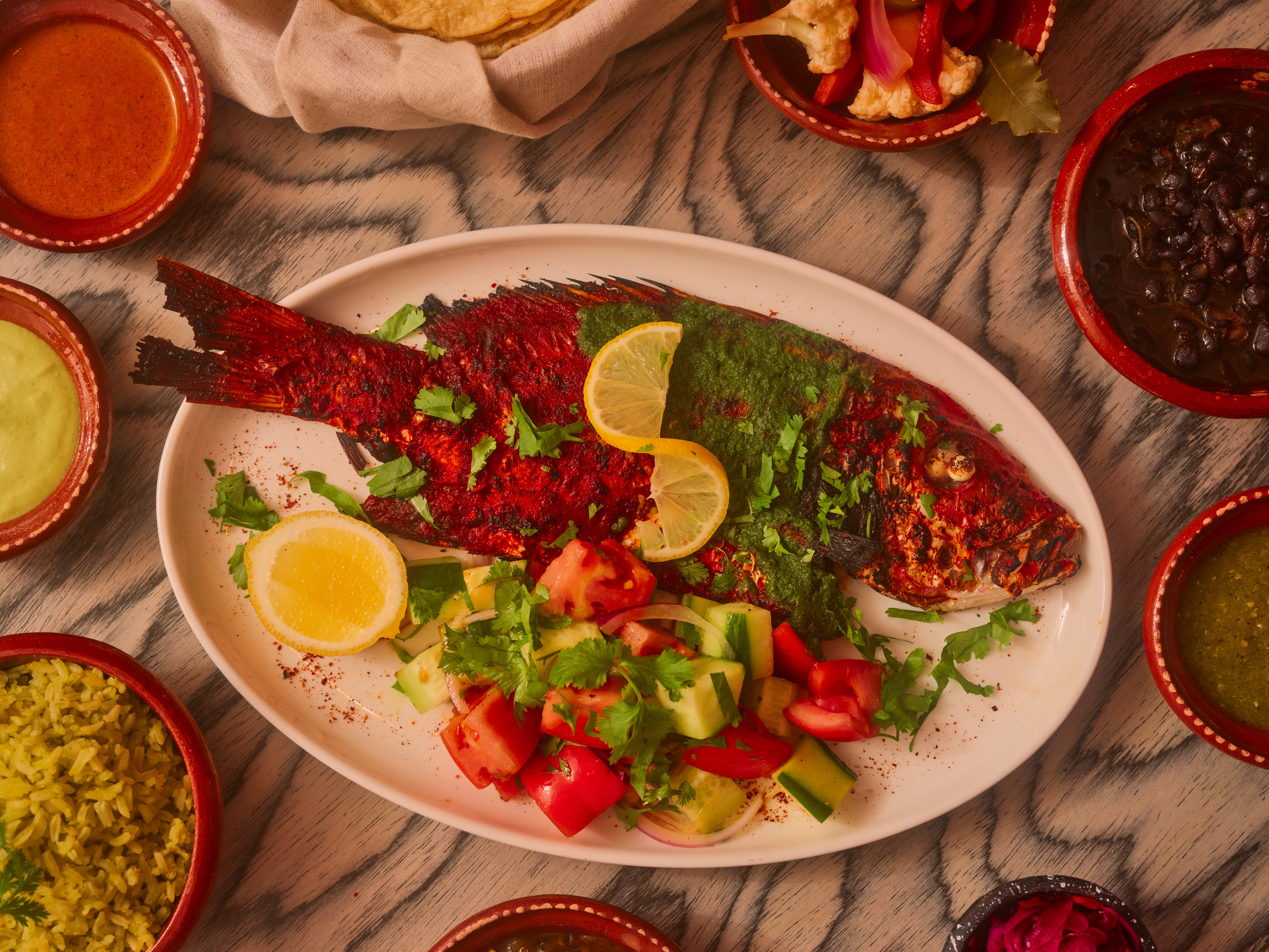 Whole fish with lime, tomatoes on a white plate on a table surrounded by sides