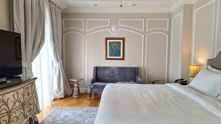 An interior shot of the King George Hotel, Athens, showing a bed, chandelier, renaissance-style aesthetic and a more modern grey sofa,