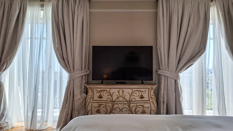 An interior view of a room at the King George Hotel Athens, taken on the bed, looking at the television on an ornate dresser, with two windows letting light in onto the wooden floor.