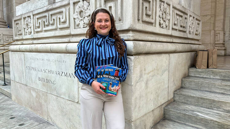 A woman in a blue shirt holds books in front of the library.