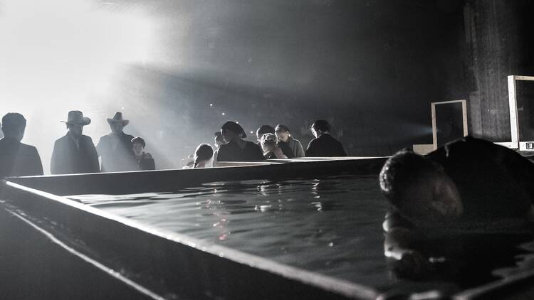 A dark scene during an opera with one person in water an others looking on.