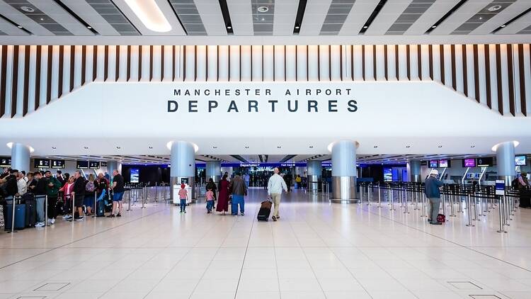 Travellers wheel suitcases towards the Departures area inside Manchester Airport which is marked by a sign above them.