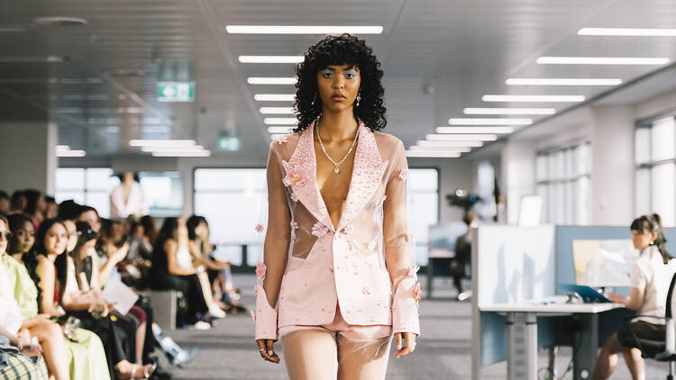 A model in a pink suit walking down a runway in an office setting.